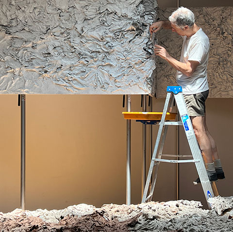 Ruggero Vanni on a stepladder in his studio, pressing a dry cast relief sheet onto a large vertical panel, the surface texture of The Wind Blowing Over the Sea visible, 2024.