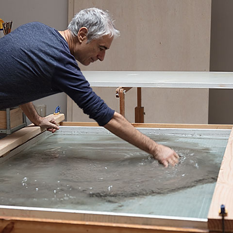 Ruggero Vanni leaning over a large studio vat, hands in the pulp suspension used to form the base sheets for The Wind Blowing Over the Sea, Brooklyn commission, 2024.