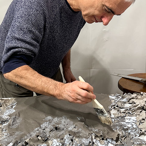 Ruggero Vanni working a wide brush over a wet pulp sheet pressed onto the relief mold, coaxing the material into the cast surface of a panel for The Wind Blowing Over the Sea, 2024.
