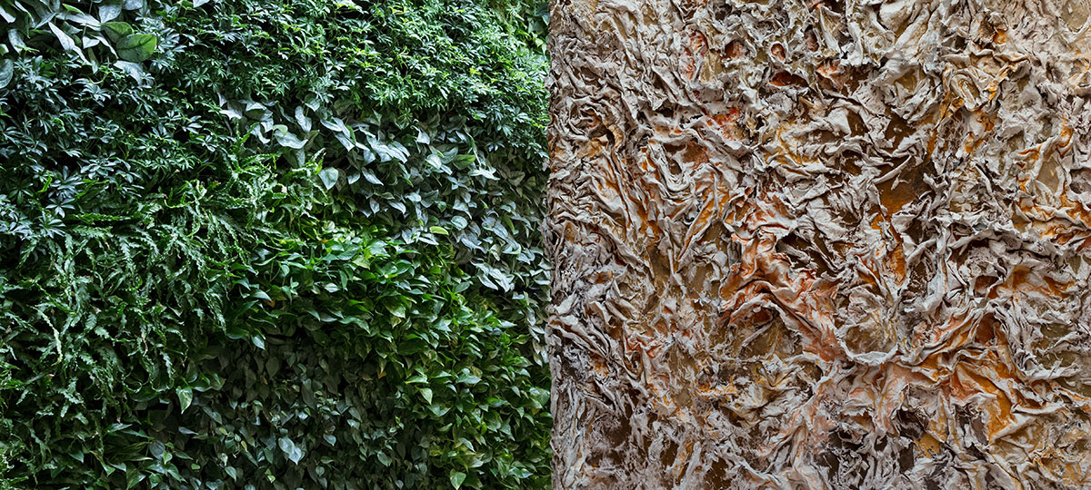 Split view contrasting the living green wall and the surface of The Wind Blowing Over the Sea by Ruggero Vanni: the deep forest and chartreuse greens of the planted wall face the earth-pigmented cast relief across the lobby of a Brooklyn residential building.