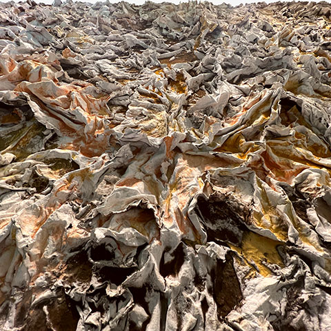 Extreme close-up of the cast recycled egg carton relief surface of The Wind Blowing Over the Sea by Ruggero Vanni, showing turbulent wave-like forms with layered earth pigments in umber, sienna, and grey, 2025.