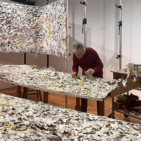 Ruggero Vanni working across multiple panels of The Wind Blowing Over the Sea laid out in sequence on studio tables, applying earth pigments to the cast relief surface, 2024.