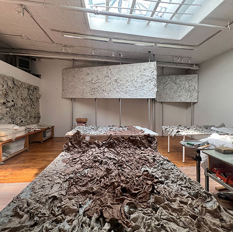 Wide view of Ruggero Vanni's studio with multiple panels of The Wind Blowing Over the Sea suspended horizontally and stacked vertically, raw cast relief surfaces visible throughout, 2024.